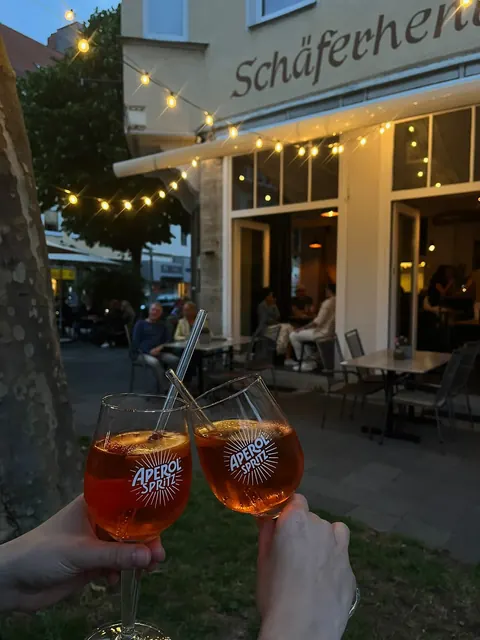 Abends auf der Terrasse des Café Heidelberg: zwei Gläser Aperol Spritz, dahinter Lichterkette, Markise und Gäste vor den erleuchteten Fenstern
