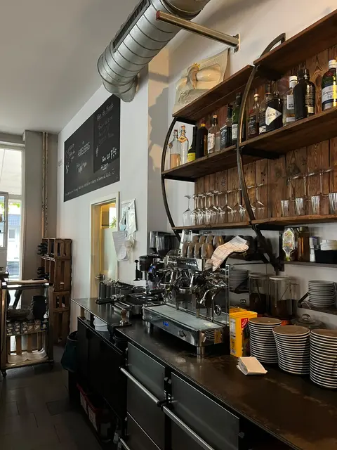 Theke mit Siebträgermaschine, Tassenstapeln und Holzregal voller Flaschen und Gläser